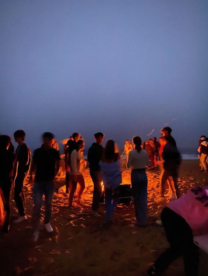 a group of people standing around a campfire on the beach at night with bright lights