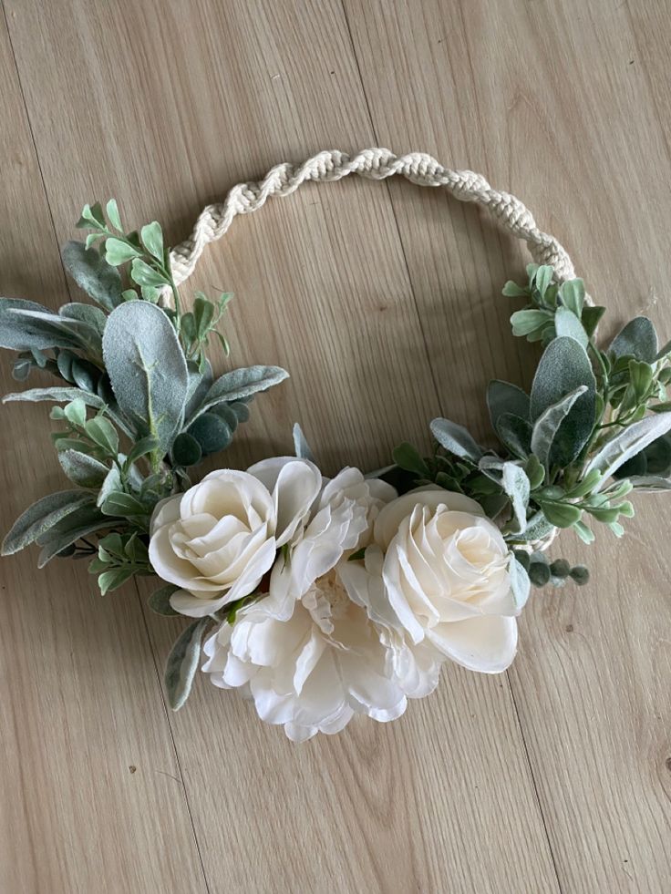 white flowers and greenery are arranged on a wooden surface in a circular wreath with rope