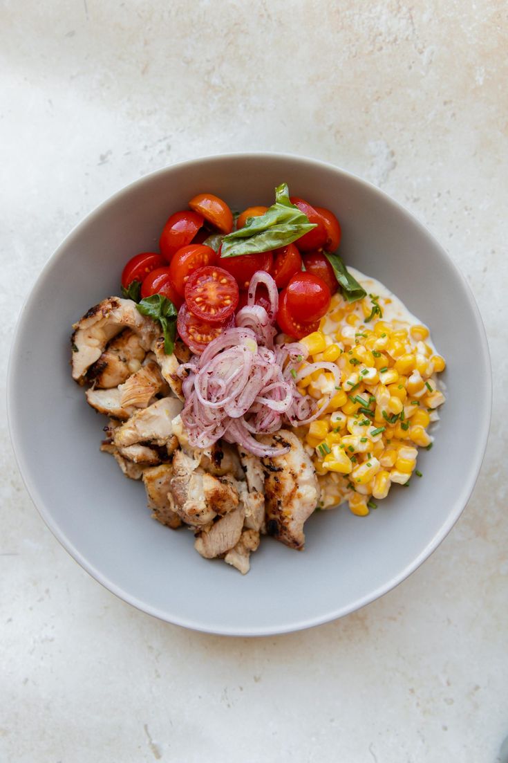 a white bowl filled with chicken, tomatoes and corn on top of a marble table