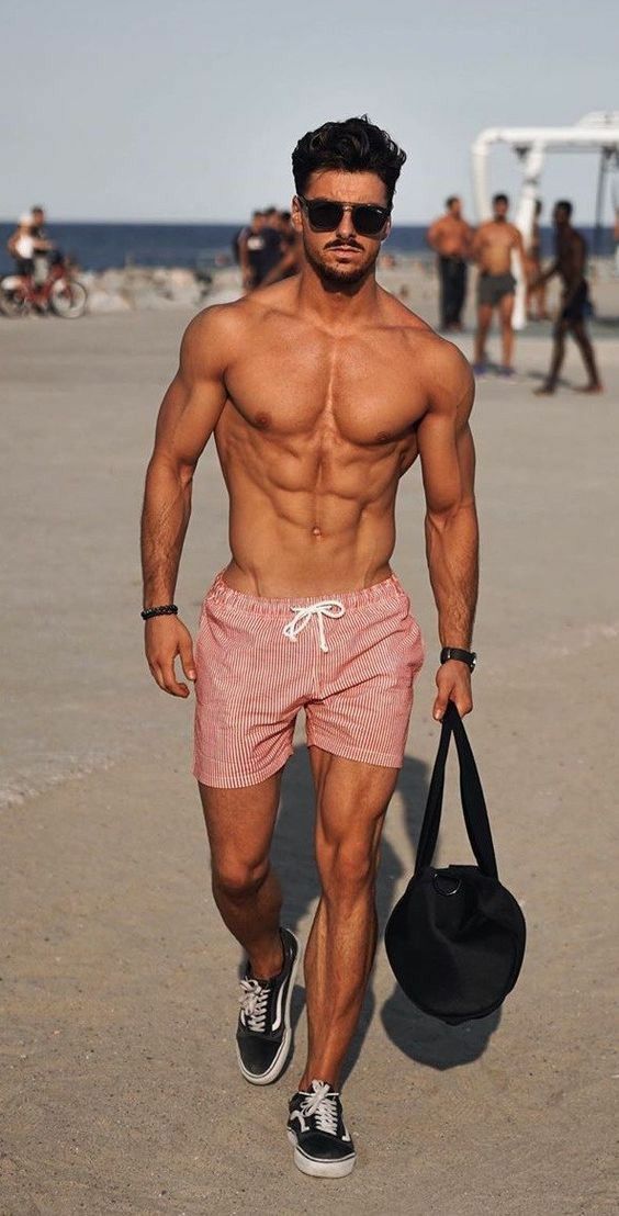 a shirtless man walking on the beach carrying a bag