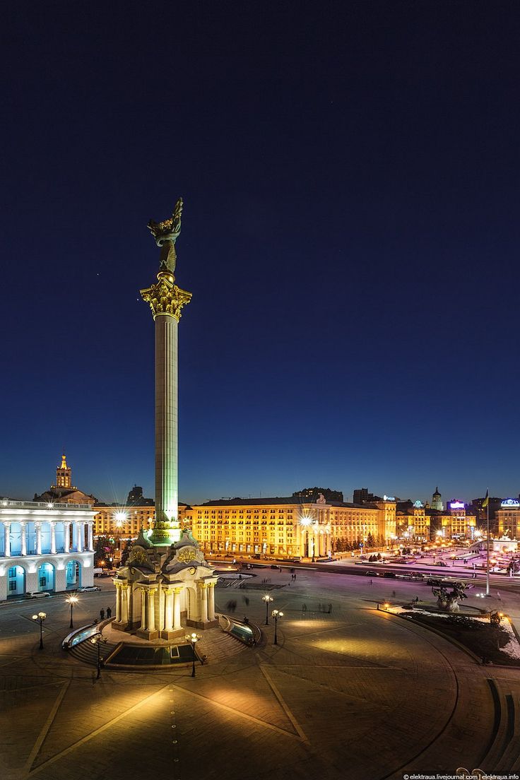 a night time view of a city square with a statue in the center and buildings around it