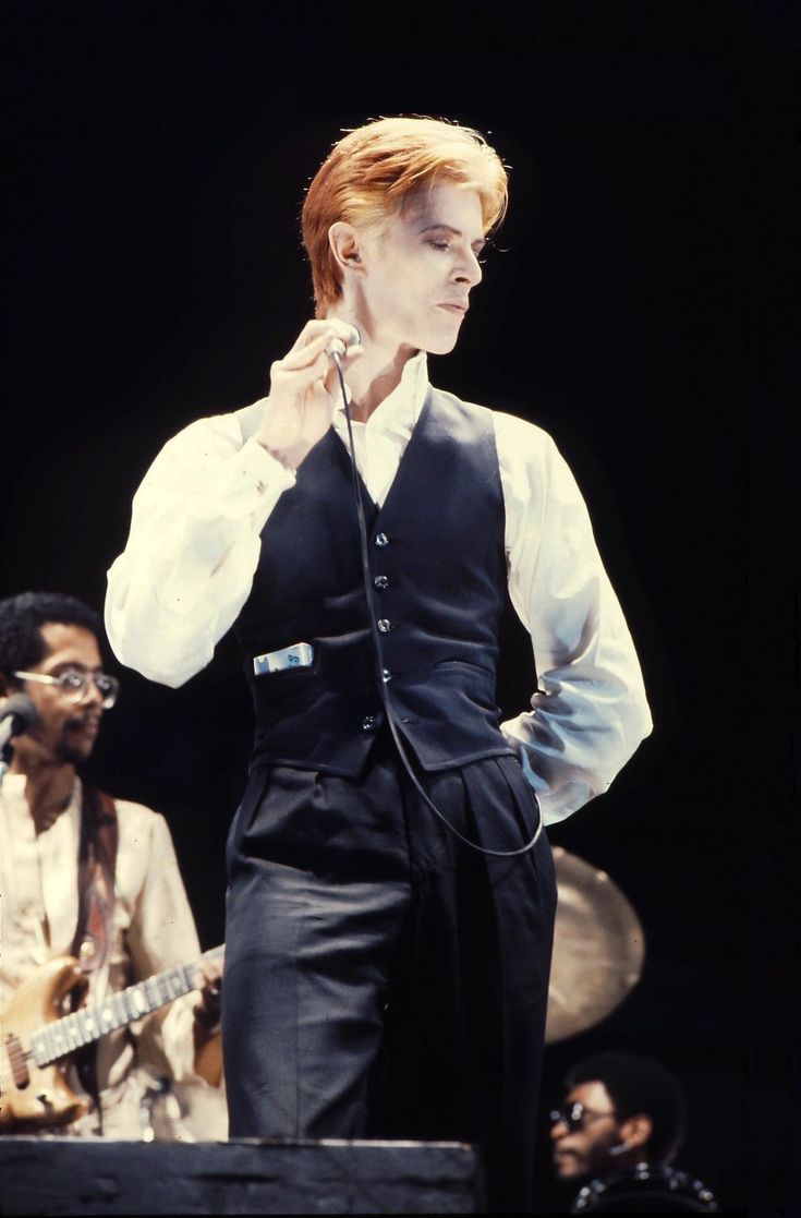 a man with red hair wearing a vest and holding a microphone in his right hand