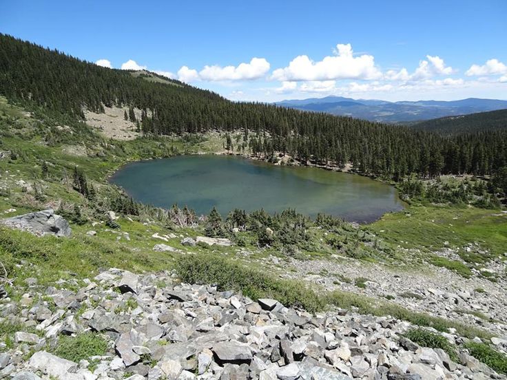 a lake surrounded by rocks and trees on the side of a hill with mountains in the background