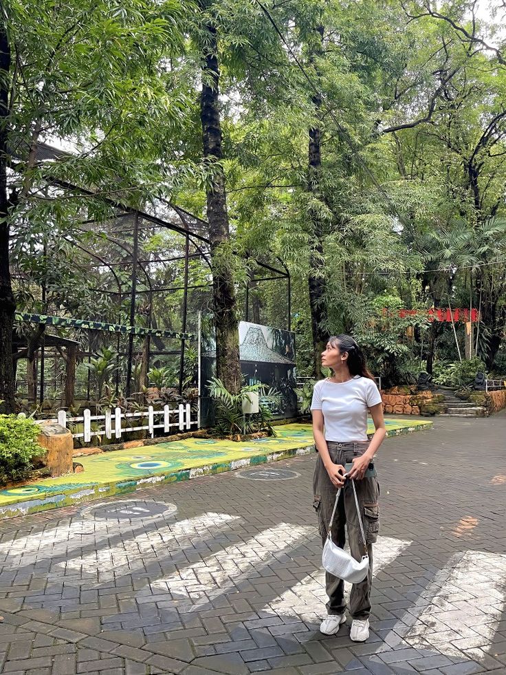 a woman standing in the middle of a park holding a white bag and looking up into the sky