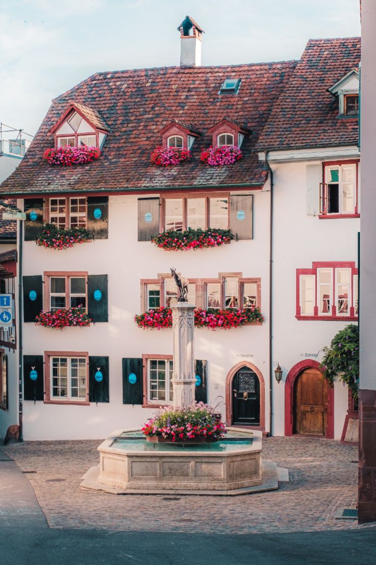 a white building with red shutters and flowers on the window sill, in front of a fountain