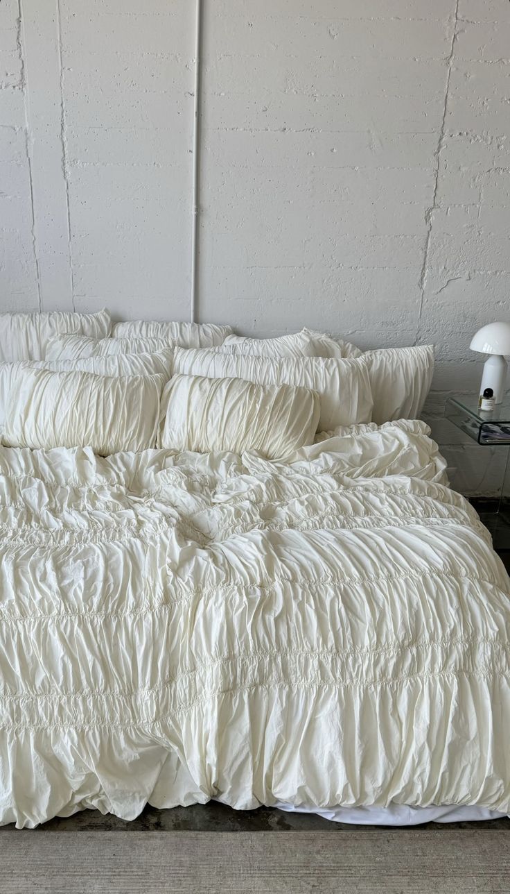 an unmade bed in front of a white brick wall and lamp on the floor