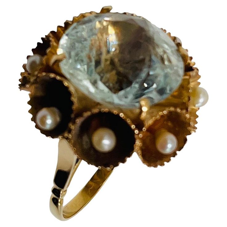 This a 14K yellow gold ring. It depicts a Brilliant Shape White Sapphire (15.18 x 15.18 x 9.73 mm ) weighing 16.14 carats and mounted on four gold prongs setting. The White Sapphire is adorned around it with a wreath made of gold trumpets with scalloped borders that are embellished with eight pearls of 2.5mm inside it. Formal Yellow Gold Rings With High Luster, High Luster Yellow Gold Rings For Formal Occasions, Formal High Luster Yellow Gold Rings, Antique Yellow Gold 14k Pearl Ring, High Luster Yellow Gold 14k Ring, Antique Round Jewelry With High Luster, Gold Topaz Ring With 17 Jewels In 14k Gold, Unique Oval Yellow Gold Topaz Ring, 14k Gold Topaz Ring With 17 Jewels