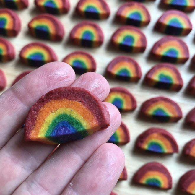 a hand holding a rainbow shaped cookie in front of rows of cookies