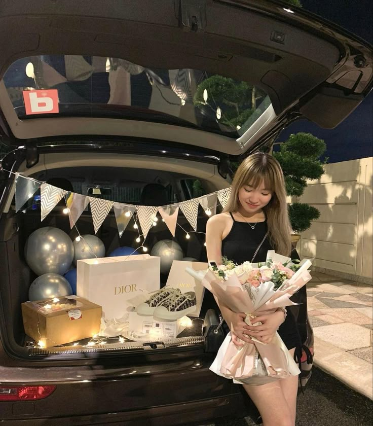 a woman standing next to the trunk of a car holding flowers and presents in front of her