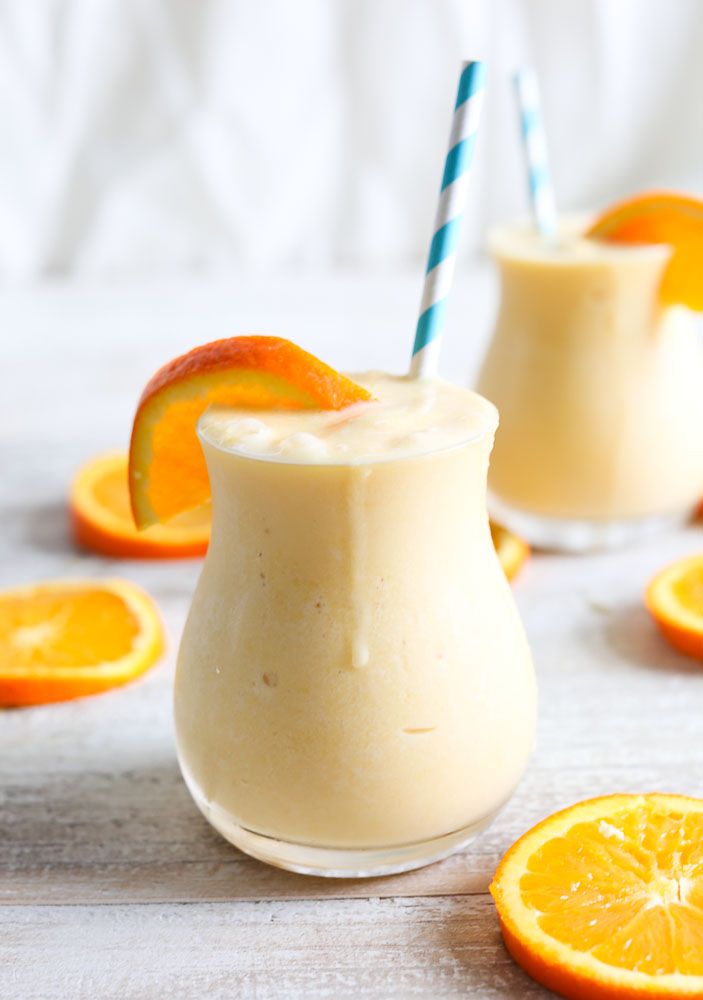 two glasses filled with orange smoothie next to sliced oranges on a white table