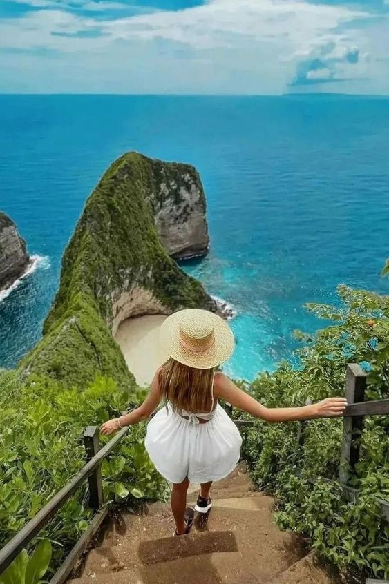 a woman in a white dress and straw hat walking down stairs towards the ocean with cliffs on either side