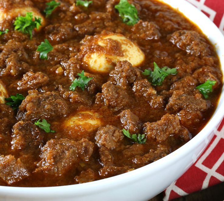 a white bowl filled with meat and potatoes on top of a red table cloth next to a wooden spoon