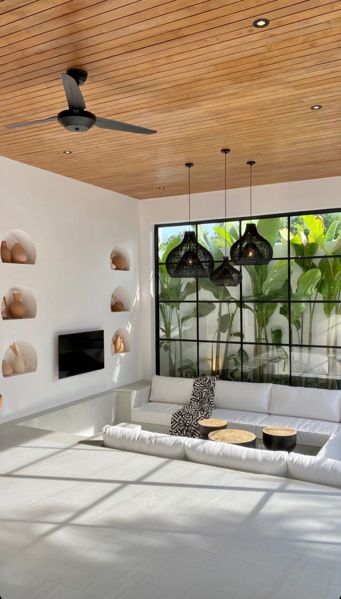 a living room filled with white furniture and lots of windows covered in plants on the wall