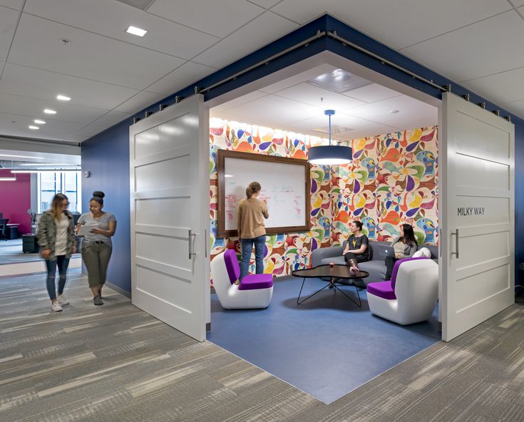 an office with people sitting in chairs and standing around the room, looking at a whiteboard