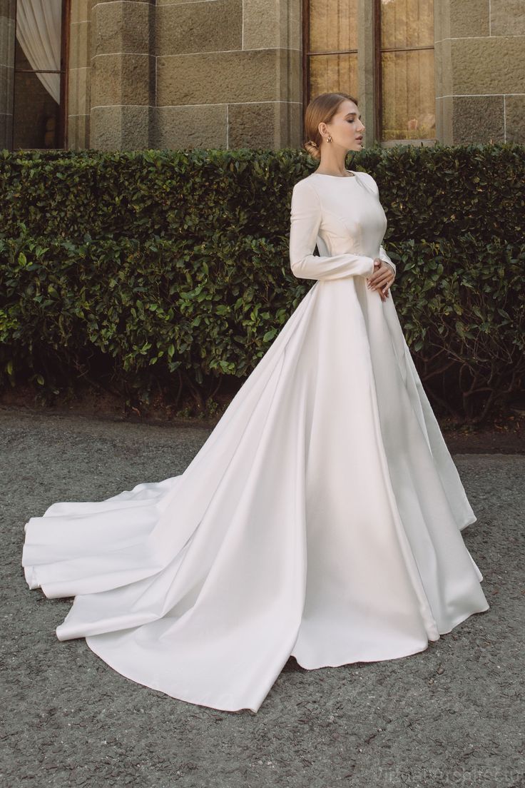 a woman standing in front of a building wearing a white wedding dress with long sleeves