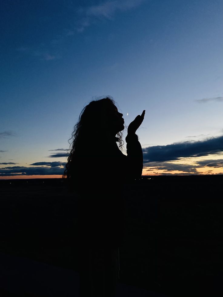 a woman is standing in the dark with her hand up to her face as the sun sets