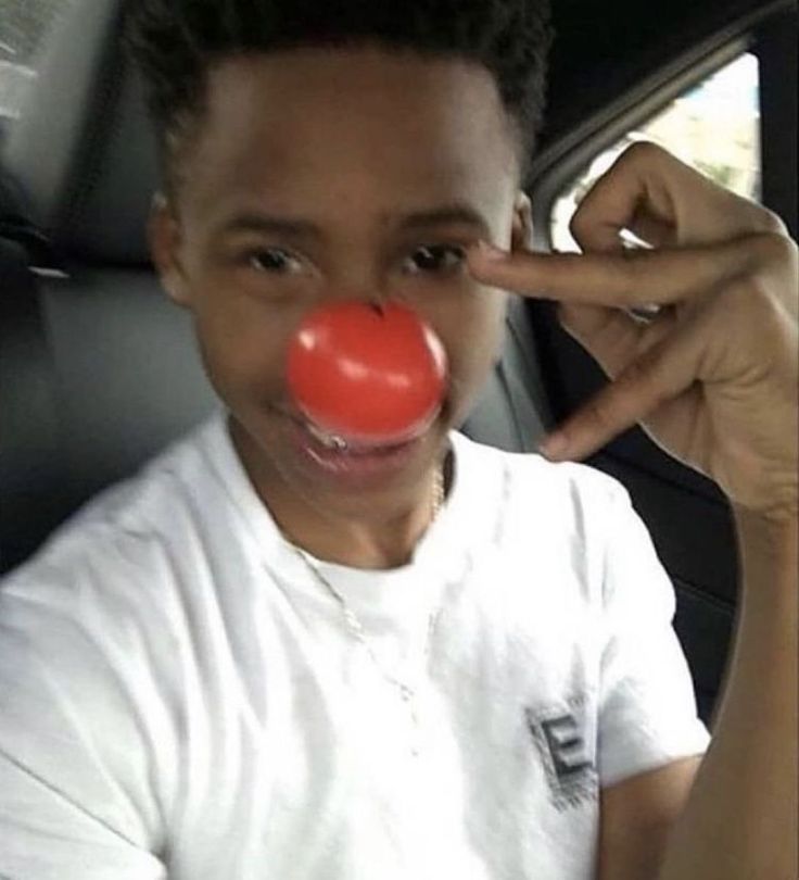 a young boy in a car with a red clown nose on his nose and one finger pointing at the camera