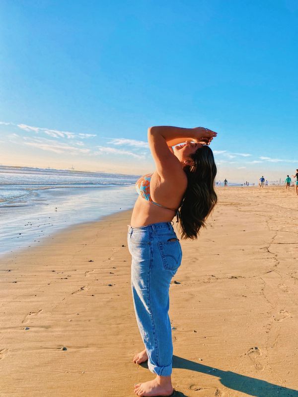 a woman standing on top of a sandy beach next to the ocean with her hands behind her head