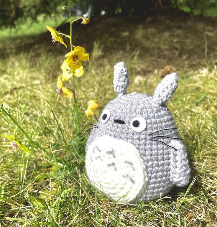 a crocheted totoro sitting in the grass next to a yellow flower