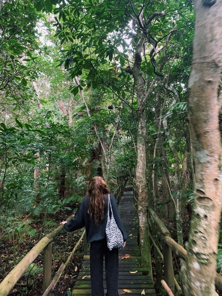 a woman walking across a wooden bridge in the middle of a forest with trees on both sides