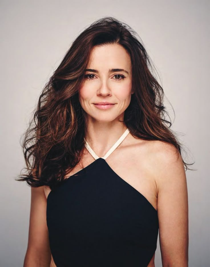 a woman with long hair wearing a black top and smiling at the camera while standing in front of a gray background