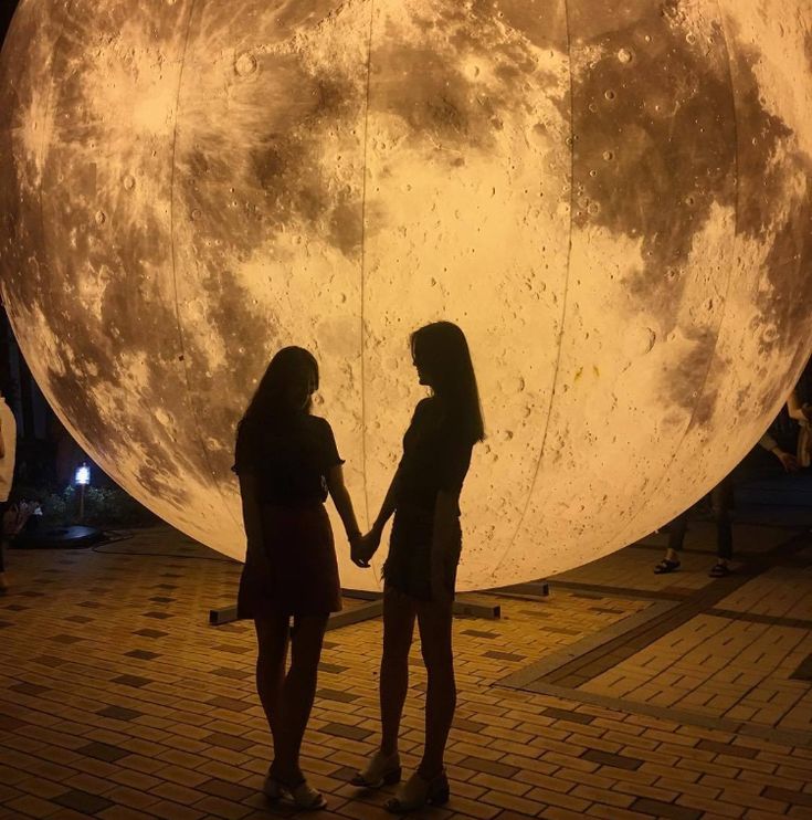 two women holding hands in front of a giant pink moon on display at the museum