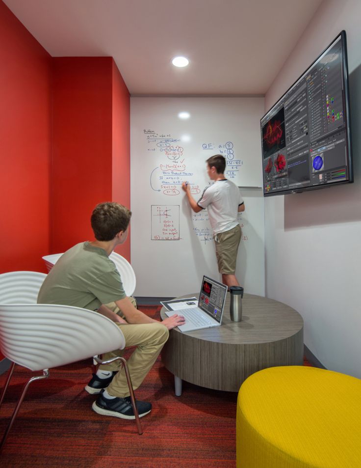 two men are working on a whiteboard in an office with red walls and chairs