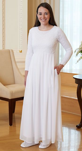 a woman standing in a room wearing a long white dress and smiling at the camera