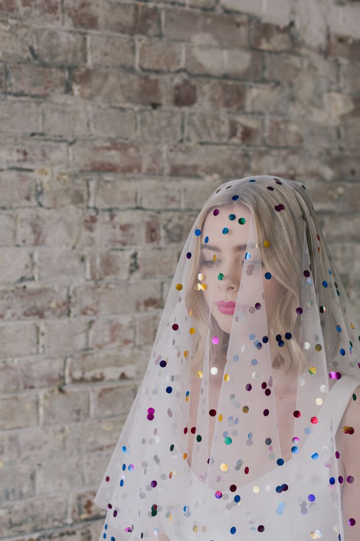 a woman wearing a veil with multicolored confetti on it and standing in front of a brick wall