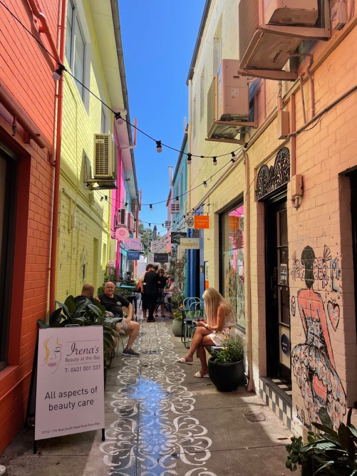 people are sitting on the sidewalk in an alleyway with colorful buildings and graffiti painted all over it