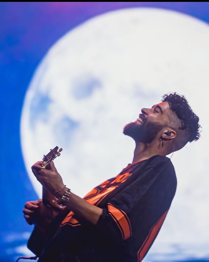 a man in an orange and black shirt singing into a microphone with the moon behind him