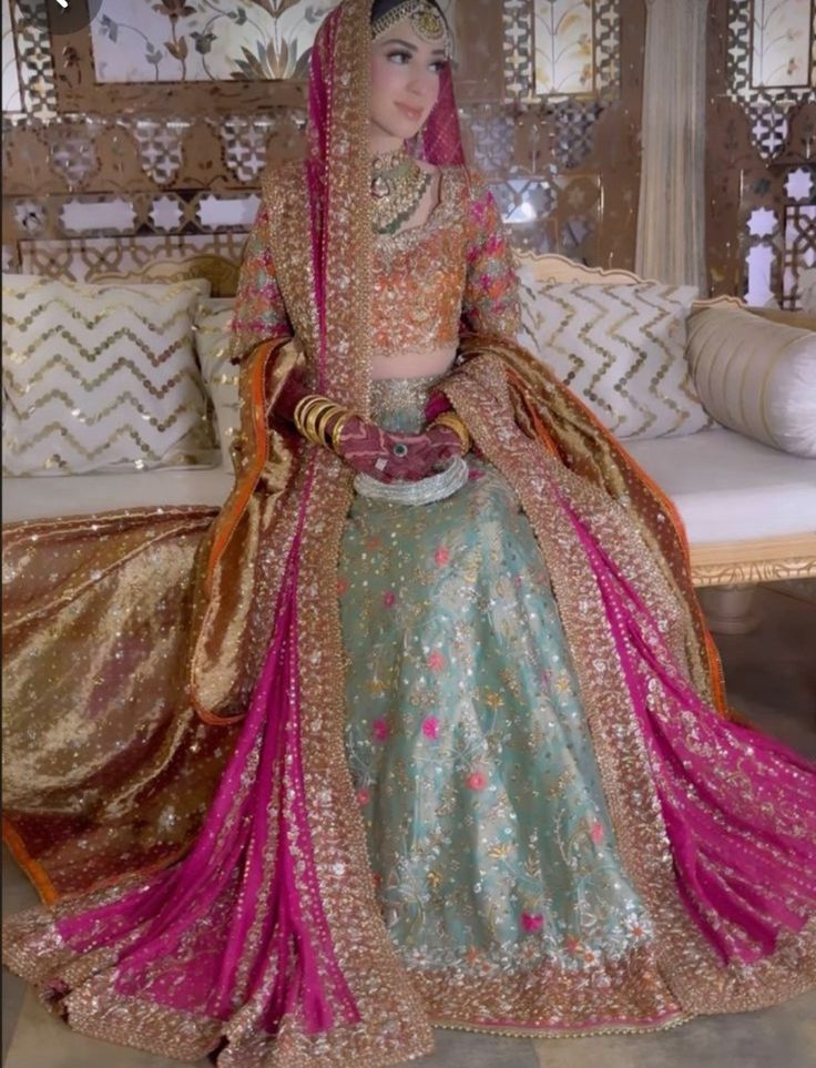 a woman in a bridal outfit sitting on a bed wearing a pink and green lehenga