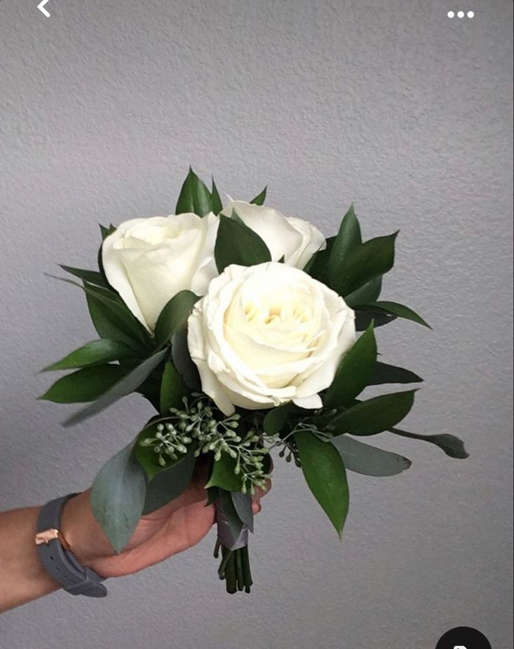 a hand holding a bouquet of white roses and greenery in front of a gray wall