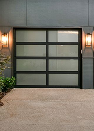 a garage door with two lights on each side and plants in the front yard next to it