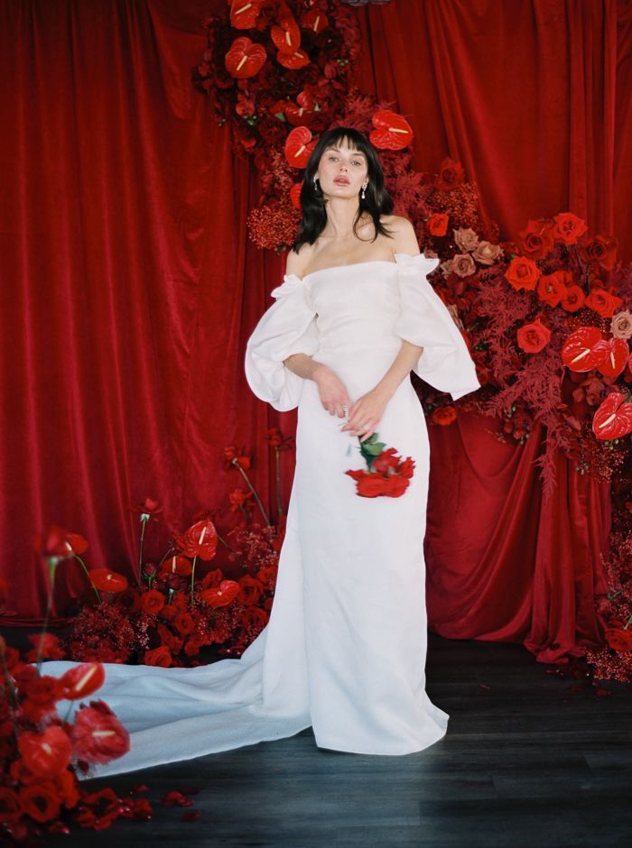 a woman standing in front of red roses wearing a white dress and holding a bouquet