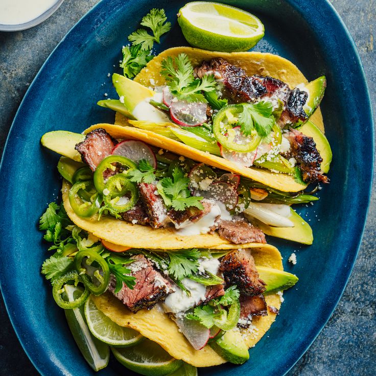 three tacos on a blue plate with limes and cilantro wedges