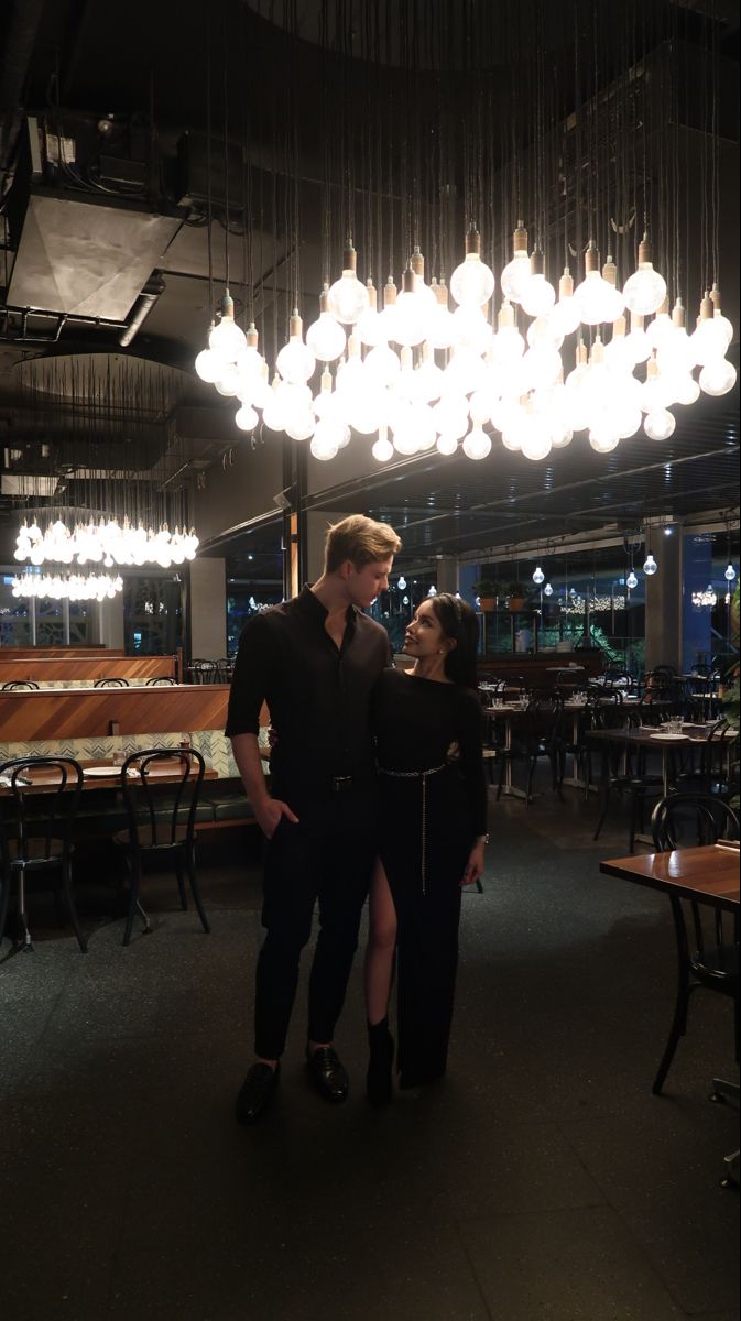 a man and woman standing under a chandelier in a room with wooden tables