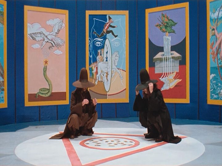 two women sitting on the floor in front of some art work and one is wearing a hat