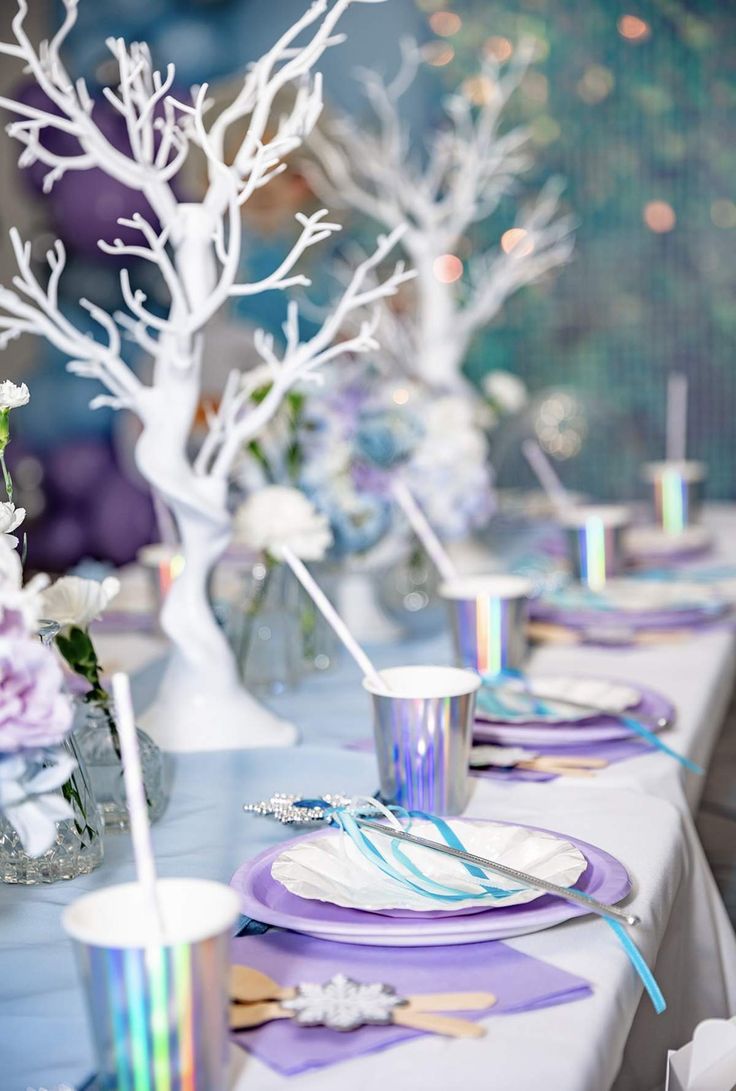 the table is set with purple and white plates, silver cups, and napkins