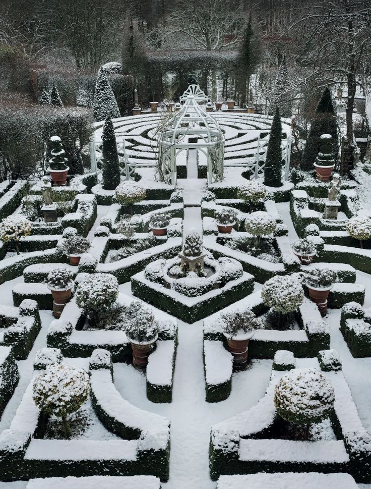 a snow covered formal garden with hedges and potted plants in the center, surrounded by trees