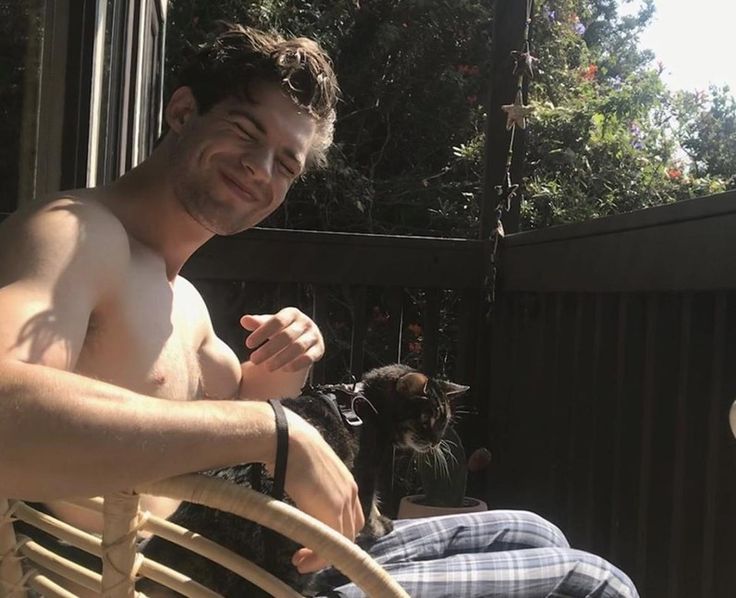 a shirtless man sitting on a wooden chair next to a cat