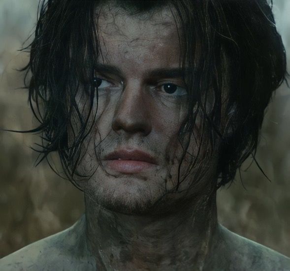 a man with wet hair and blue eyes stares into the camera while standing in front of some grass