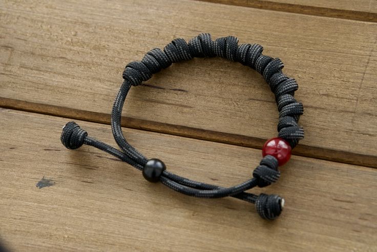 This stunning accessory is designed to help you stay close to our Lord while wearing your faith on your sleeve. Crafted with durable, premium paracord rope in a sleek black/gray color, this adjustable bracelet features 10 knots that represent the 10 Hail Mary prayers. But what truly sets it apart is its unique milky red Our Father bead, reminiscent of the fiery passion and devotion required to vanquish the forces of darkness. Wear this bracelet as a powerful reminder of your commitment to prayer Black Beaded Bracelet With Sliding Knot, Black Spiritual Friendship Bracelets With Adjustable Cord, Black Spiritual Friendship Bracelet With Adjustable Cord, Spiritual Black Adjustable Friendship Bracelets, Spiritual Black Friendship Bracelets With Adjustable Cord, Spiritual Black Braided Bracelets For Friendship, Casual Black Beaded Bracelets For Friendship, Black Braided Casual Friendship Bracelets, Black Spiritual Friendship Bracelets With Sliding Knot