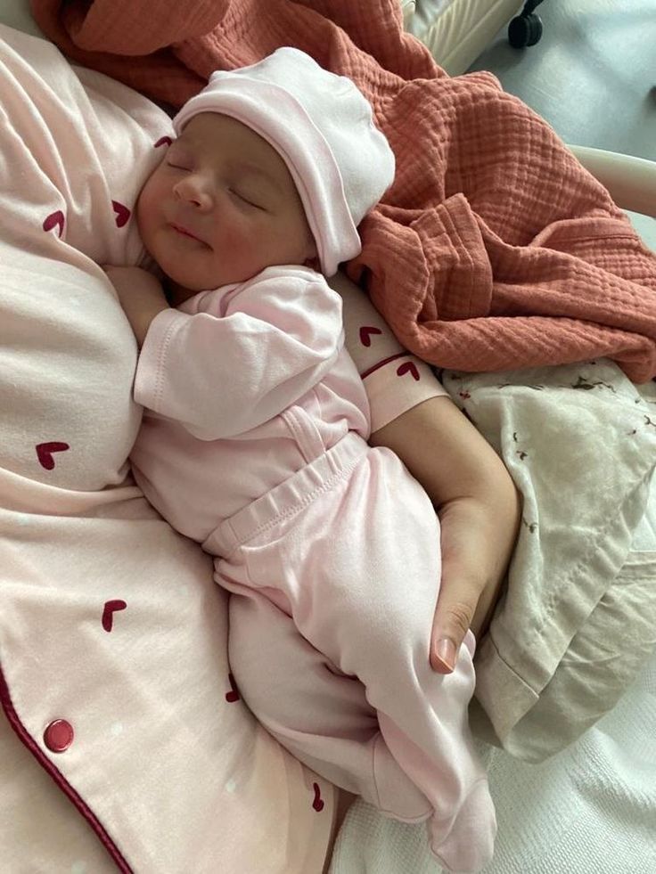 a baby sleeping in a crib next to a teddy bear wearing a pink hat