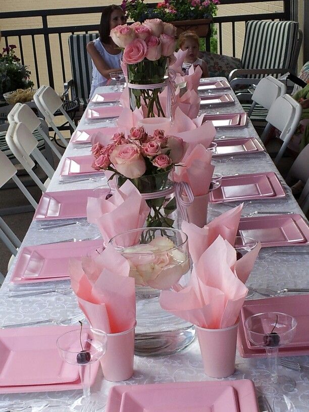 a long table with pink napkins and flowers in vases on the top, along with other place settings