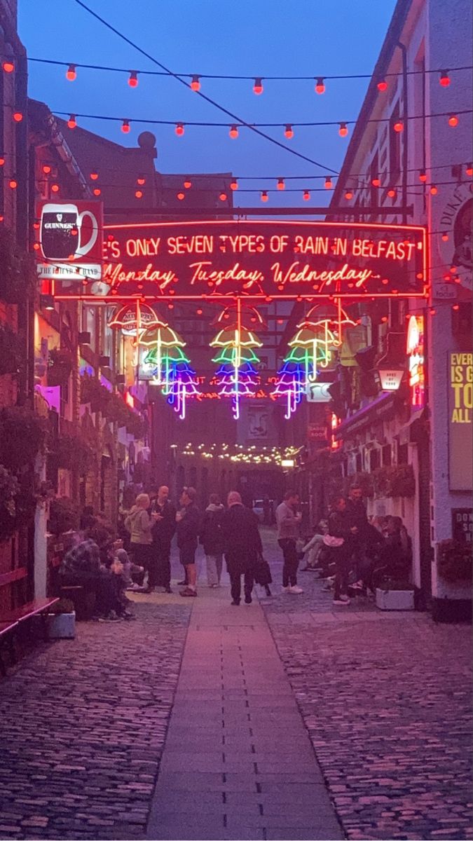 people are walking down an alley way with lights strung over the buildings and signs above them