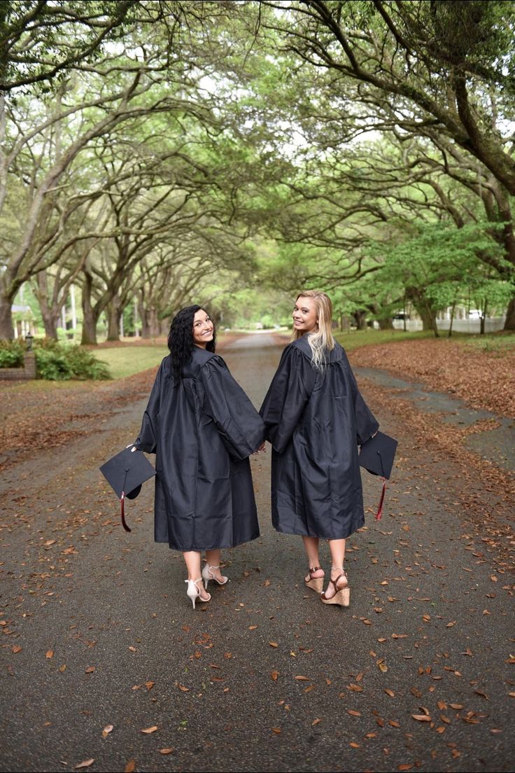 two women in graduation gowns walking down the road together with trees lining both sides