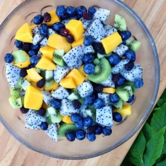 a glass bowl filled with fruit on top of a wooden cutting board next to green leaves