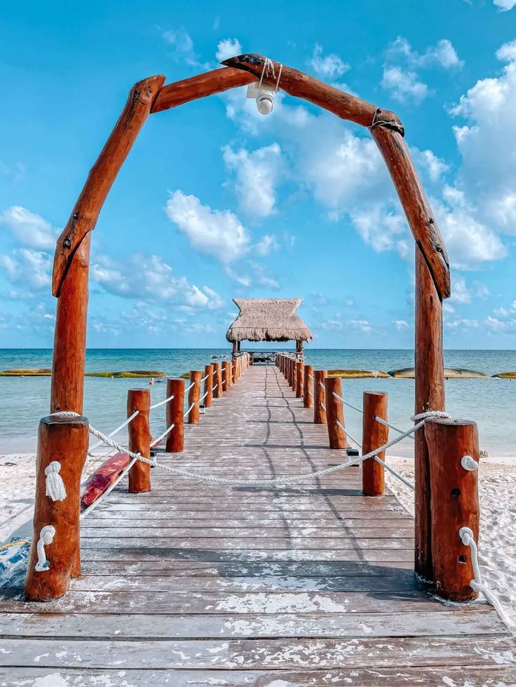 a wooden pier on the beach with a thatched hut in the background and blue sky