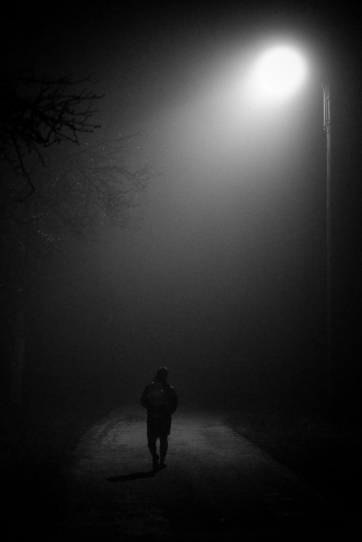 a person walking down a dark road at night with street lights in the fog behind them